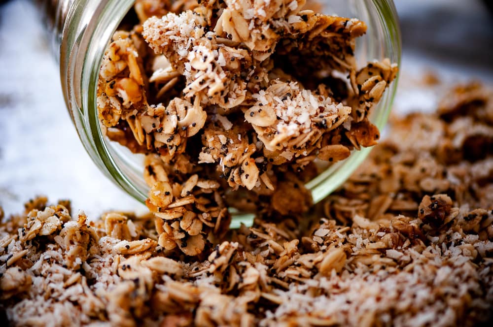 Loose granola with oats and coconut flakes spills from a glass jar onto a surface, perfect for topping your favorite fresh vegan recipes.