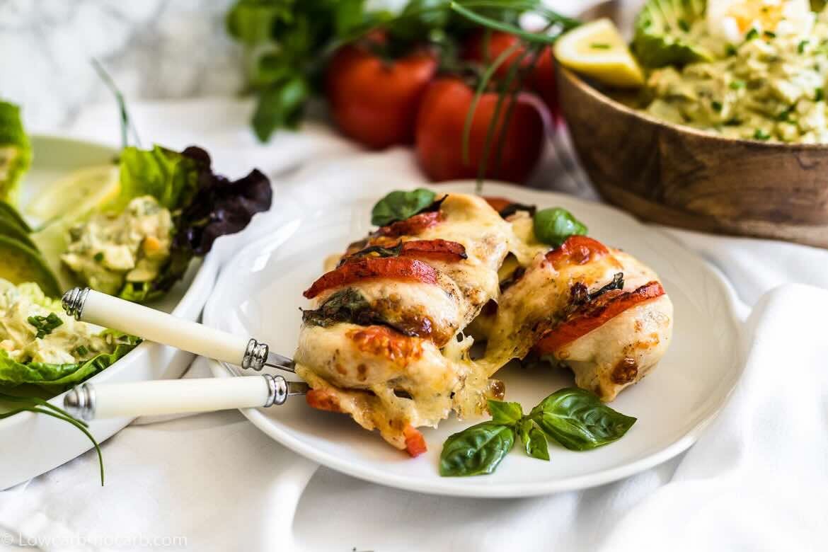 A plate with a stuffed chicken breast topped with melted cheese, tomatoes, and herbs, garnished with fresh basil—an easy dinner idea—with salad and cutlery beside it.