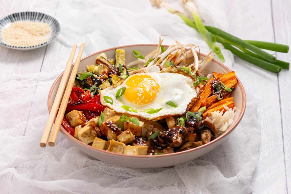 A bowl of rice topped with tofu, vegetables, a sunny-side-up egg, scallions, and sauce—easy dinners like this are perfect for busy nights, with chopsticks resting on the side.