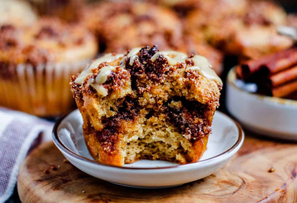 Close-up of a cinnamon muffin with a bite taken out, showing a swirl of cinnamon filling and glaze on top, placed on a small plate—perfect for cozy bakes or breakfasts with cinnamon. Other muffins appear in the background.