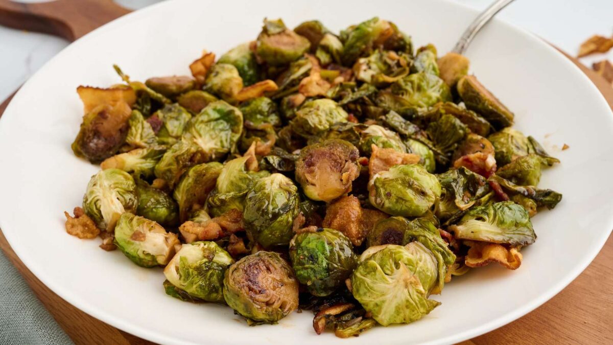 A white plate filled with roasted Brussels sprouts and crispy bacon pieces, showcasing one of the tastiest Brussels Sprouts dishes, with a fork partially visible at the edge of the plate.