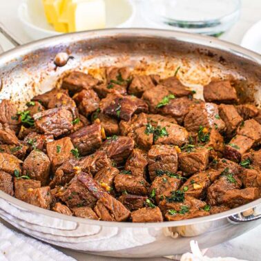 A stainless steel pan filled with cooked beef steak bites, garnished with herbs, sits on a counter near a plate, butter, and seasonings—perfect for easy dinners or quick weeknight dinners.