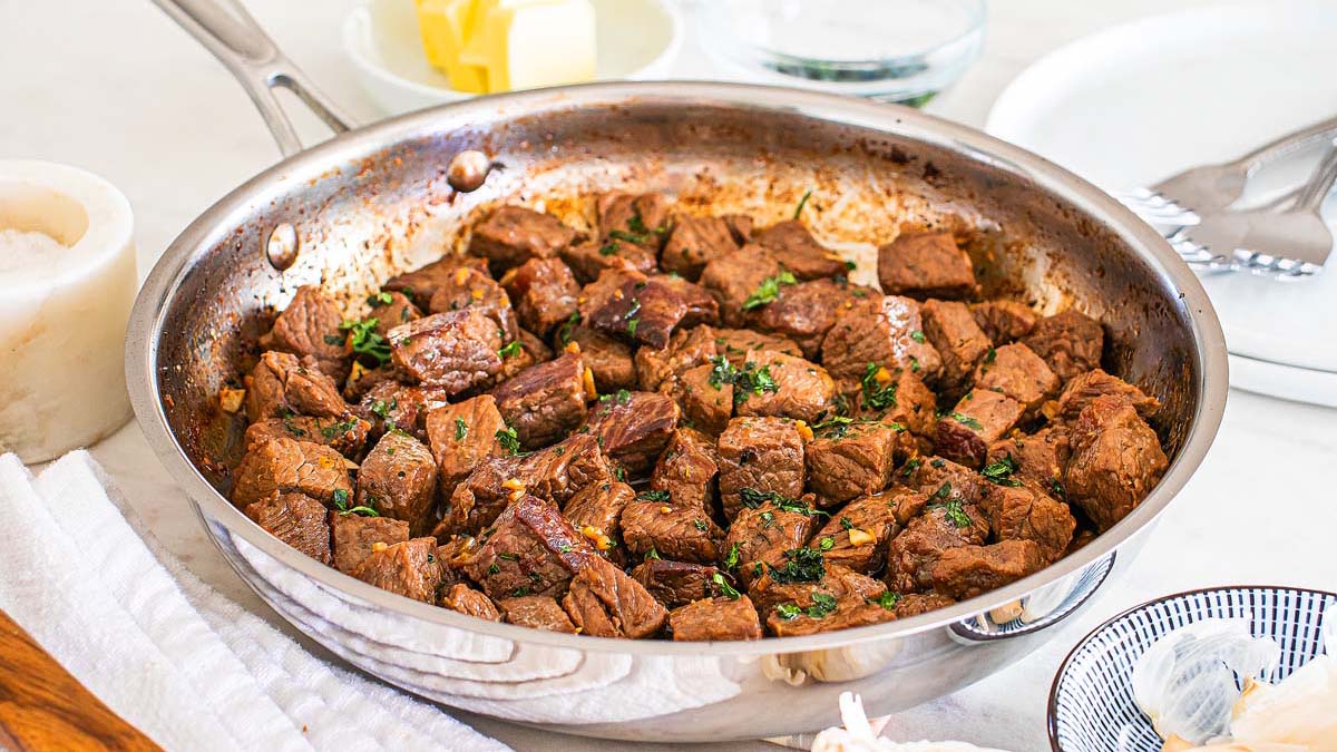 A stainless steel pan filled with cooked beef steak bites, garnished with herbs, sits on a counter near a plate, butter, and seasonings—perfect for easy dinners or quick weeknight dinners.