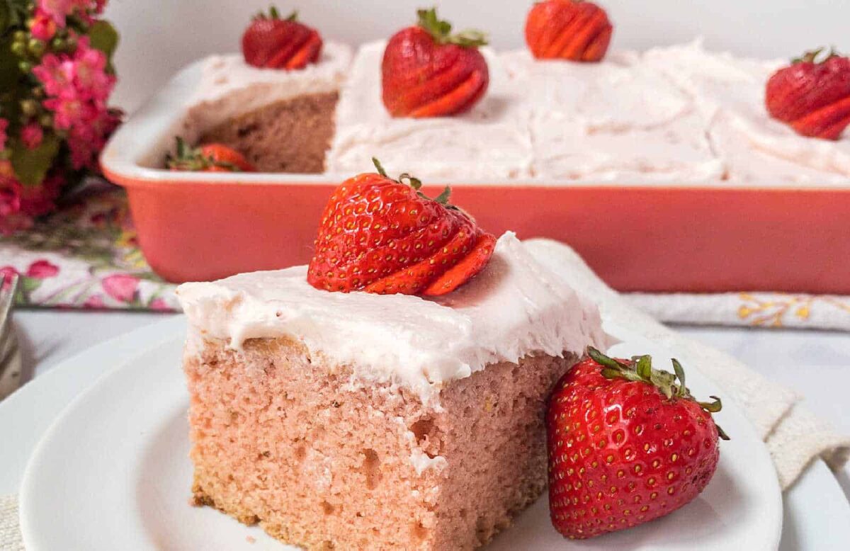 A slice of frosted strawberry cake, perfect for morning cakes, is topped with fresh strawberries on a white plate, while the rest rests in a pink baking dish in the background.