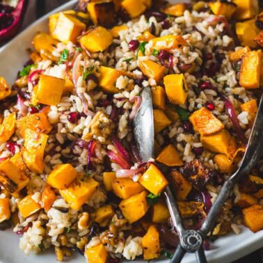 A serving platter with a vibrant mixed rice salad, roasted butternut squash cubes, red onions, walnuts, and fresh herbs—showcasing how rice bowl recipes can come together easily with simple ingredients. Includes a pair of serving tongs.