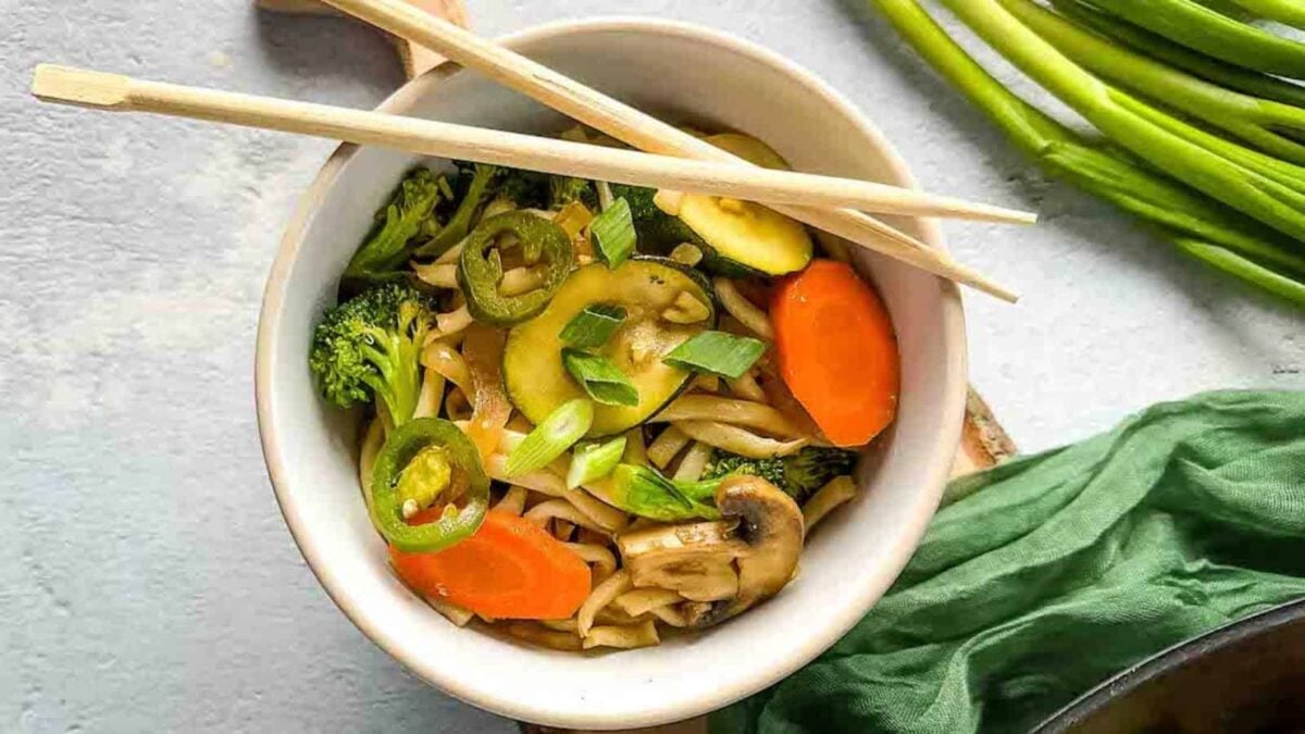 A bowl of vegetable stir-fry noodles with mushrooms, carrots, broccoli, zucchini, green onions, and jalapeños—perfect for quick meals or easy weeknight dinners—with wooden chopsticks resting on the bowl.