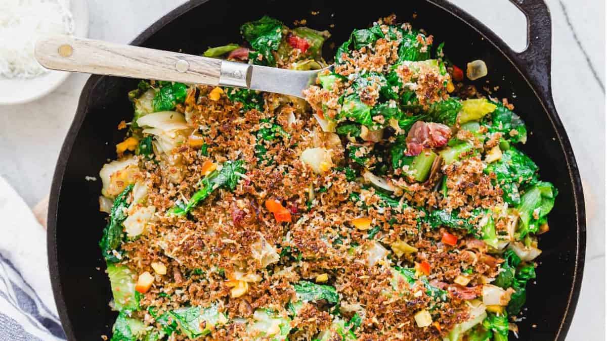 A cast iron skillet filled with a baked vegetable casserole topped with golden toasted breadcrumbs, making it perfect for side dishes or as a hearty main course, with a serving spoon placed inside.