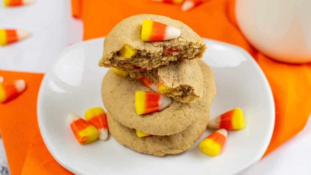 Three cookies topped with candy corn are stacked on a white plate, surrounded by scattered candy corn on an orange surface—these disappearing cookies are perfect for anyone who loves quick cookie recipes.