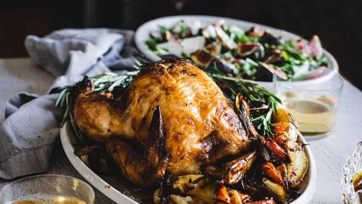 A roasted whole chicken on a platter with herbs and vegetables, paired with a glass of white wine and salad—perfect for simple dinners or easy chicken dinners at home.