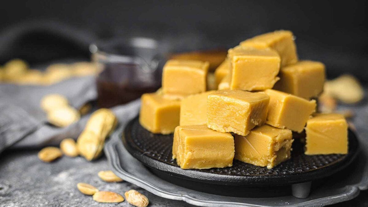 A stack of square pieces of peanut butter fudge arranged on a black plate, surrounded by peanuts and a glass cup in the background—these magical treats are perfect for anyone who loves delicious desserts.