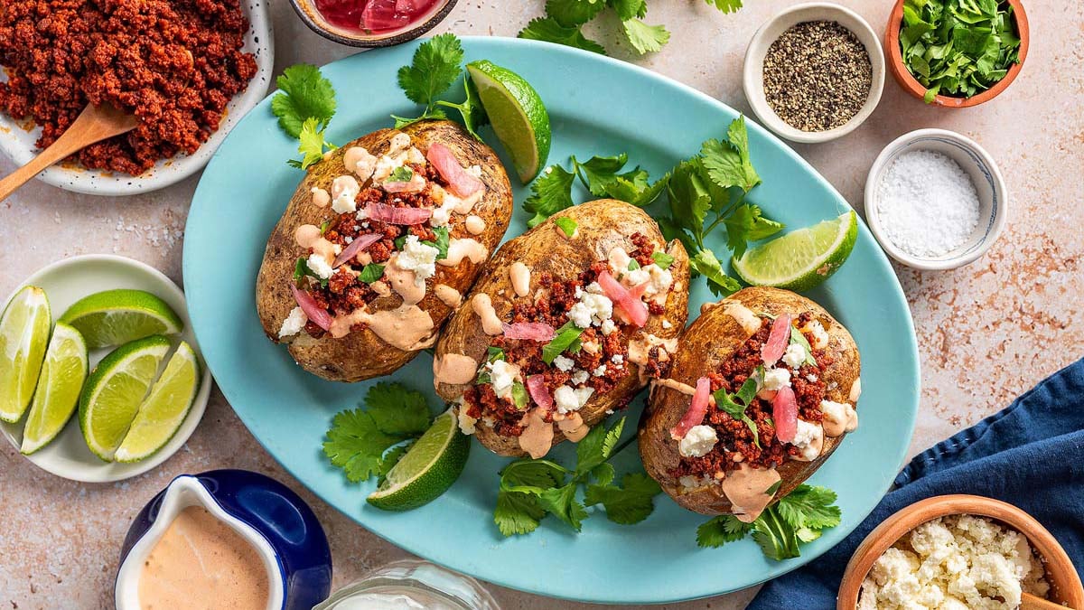 Three baked potatoes topped with crumbled meat, cheese, sauce, and diced onions are served on a blue plate with fresh cilantro and lime wedges—perfect for fresh lunch ideas with no leftover taste, surrounded by various condiments.