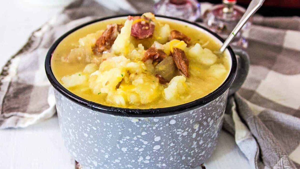 A bowl of thick, yellow split pea soup with chunks of ham and vegetables, served in a gray speckled bowl with a spoon—perfect before enjoying your favorite dairy free desserts.