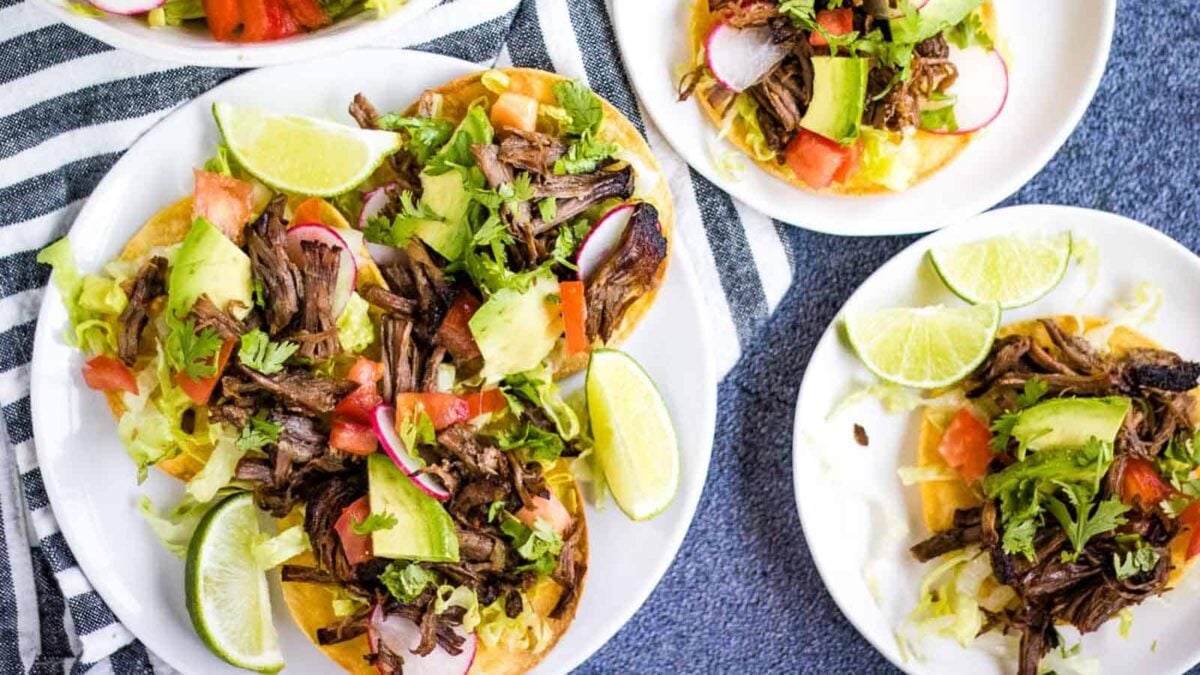 Plates of beef tostadas topped with shredded beef, lettuce, tomato, avocado, radish, cilantro, and lime wedges—one of the best beef dishes to enjoy for easy beef meals—are arranged on a striped cloth.