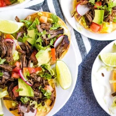 Plates of beef tostadas topped with shredded beef, lettuce, tomato, avocado, radish, cilantro, and lime wedges—one of the best beef dishes to enjoy for easy beef meals—are arranged on a striped cloth.