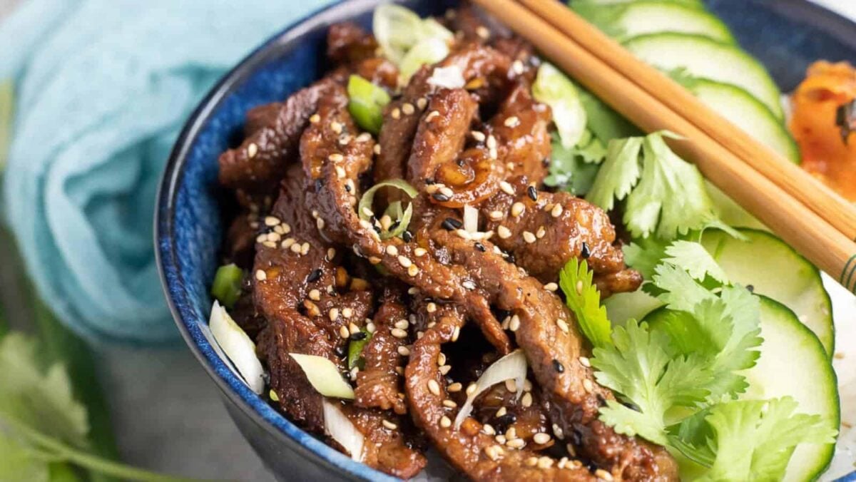 A bowl of Korean beef bulgogi, one of the most flavorful beef dishes, topped with sesame seeds, sliced green onions, cilantro, and cucumber, with chopsticks resting on the bowl.