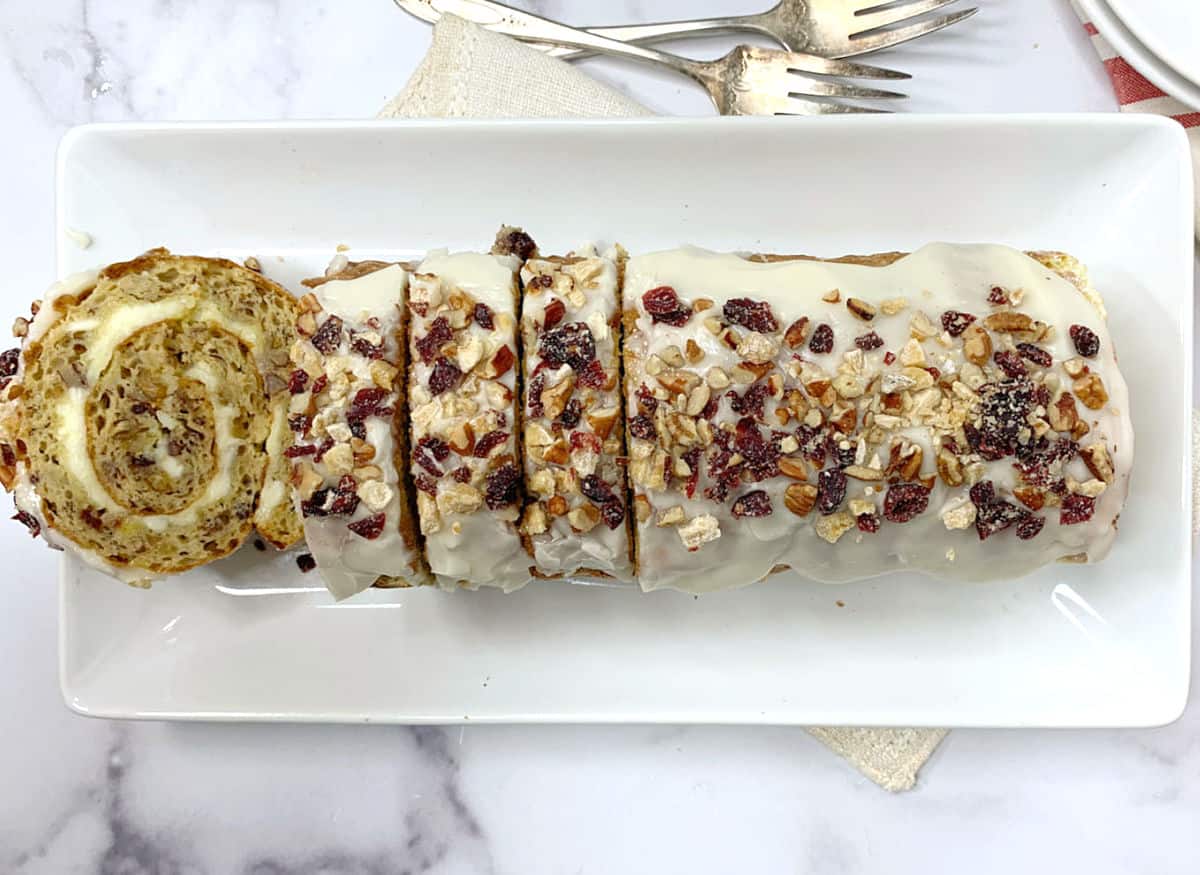 A sliced, frosted rolled cake—one of our best easy cakes—topped with chopped nuts and dried fruit is arranged on a rectangular white plate, with dessert forks in the background.