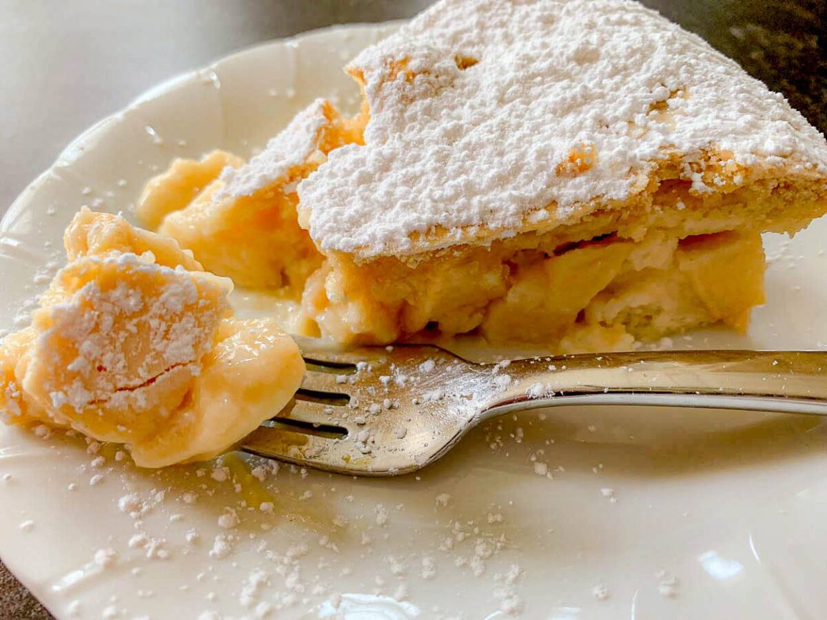 A slice of homemade custard pie topped with powdered sugar is served on a white plate, with a fork holding a bite-sized piece—perfect for company or those who love cakes.