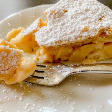 A slice of homemade custard pie topped with powdered sugar is served on a white plate, with a fork holding a bite-sized piece—perfect for company or those who love cakes.