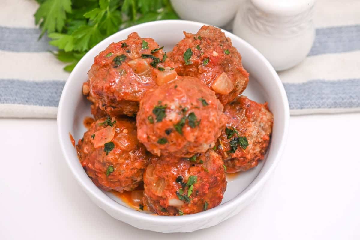 A white bowl filled with six meatballs in tomato sauce, garnished with chopped parsley, showcases one of the best easy pork recipes. It sits on a white surface near a striped cloth and fresh parsley leaves.