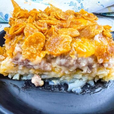 A slice of casserole with a crispy cornflake topping, ground meat layer, creamy sauce, and a base of shredded potatoes on a black plate—perfect for hearty breakfasts or adding to your collection of breakfast recipes.