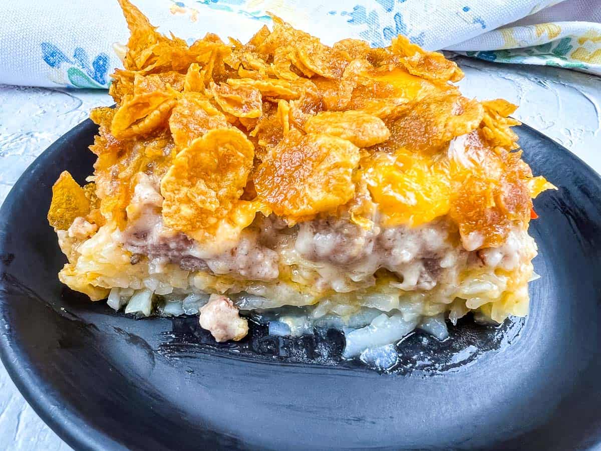 A slice of casserole with a crispy cornflake topping, ground meat layer, creamy sauce, and a base of shredded potatoes on a black plate—perfect for hearty breakfasts or adding to your collection of breakfast recipes.