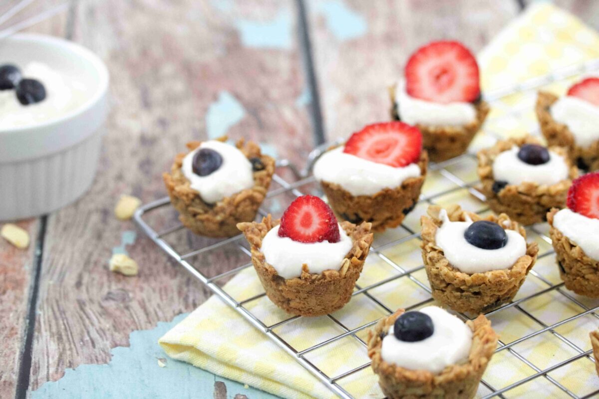 Mini granola cups filled with yogurt and topped with strawberry or blueberry halves make the best snacks, displayed on a cooling rack with a yellow cloth and a bowl of yogurt on the side.
