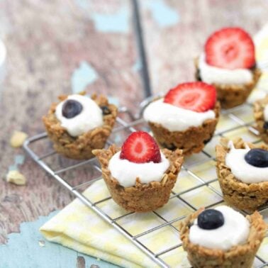 Mini granola cups filled with yogurt and topped with strawberry or blueberry halves make the best snacks, displayed on a cooling rack with a yellow cloth and a bowl of yogurt on the side.