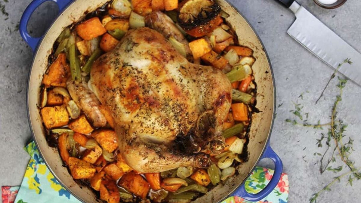 A roasted whole chicken seasoned with herbs, surrounded by sweet potatoes, celery, and onions in a blue Dutch oven—perfect for those seeking fast dinners with hearty flavors and minimal prep.