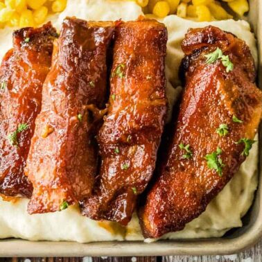 A serving of slow cooker pork ribs glazed to perfection on mashed potatoes, with a side of corn, garnished with parsley, in a square dish on a rustic wooden surface.