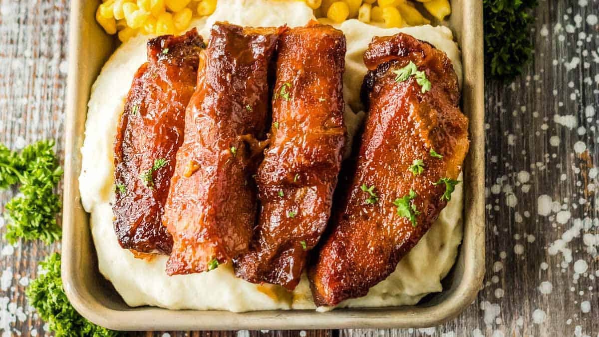 A serving of slow cooker pork ribs glazed to perfection on mashed potatoes, with a side of corn, garnished with parsley, in a square dish on a rustic wooden surface.