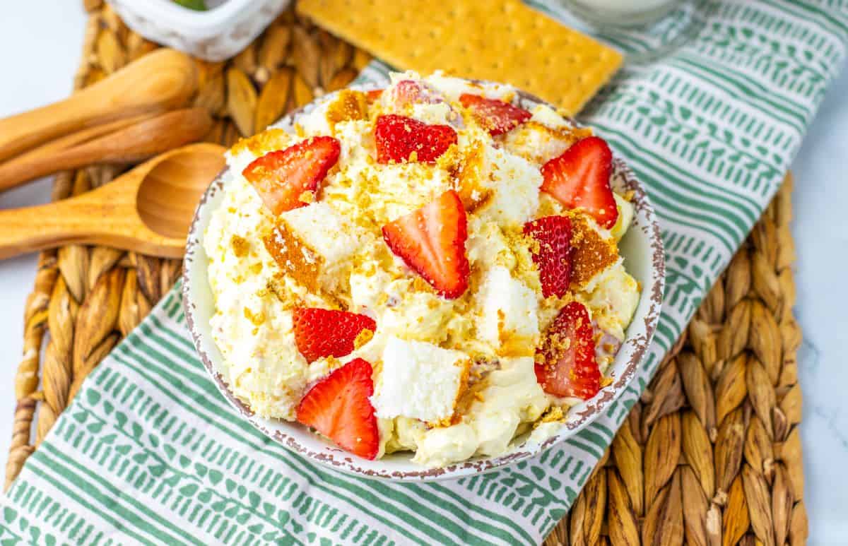 A bowl of dessert salad with whipped topping, graham cracker pieces, and sliced strawberries sits on a woven mat beside wooden spoons and a stack of graham crackers—perfect for fans of easy desserts and no bake treats.