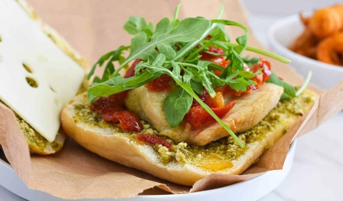 A delicious takeout alternative, this sandwich features chicken breast, sun-dried tomatoes, pesto spread, arugula, and Swiss cheese on a sub roll, served partly open-faced on brown paper—perfect for healthy lunches.