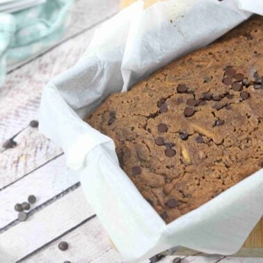 A loaf of dairy free chocolate chip banana bread in a parchment-lined baking pan on a light wooden surface—perfect for those who love indulgent lux desserts without the dairy.