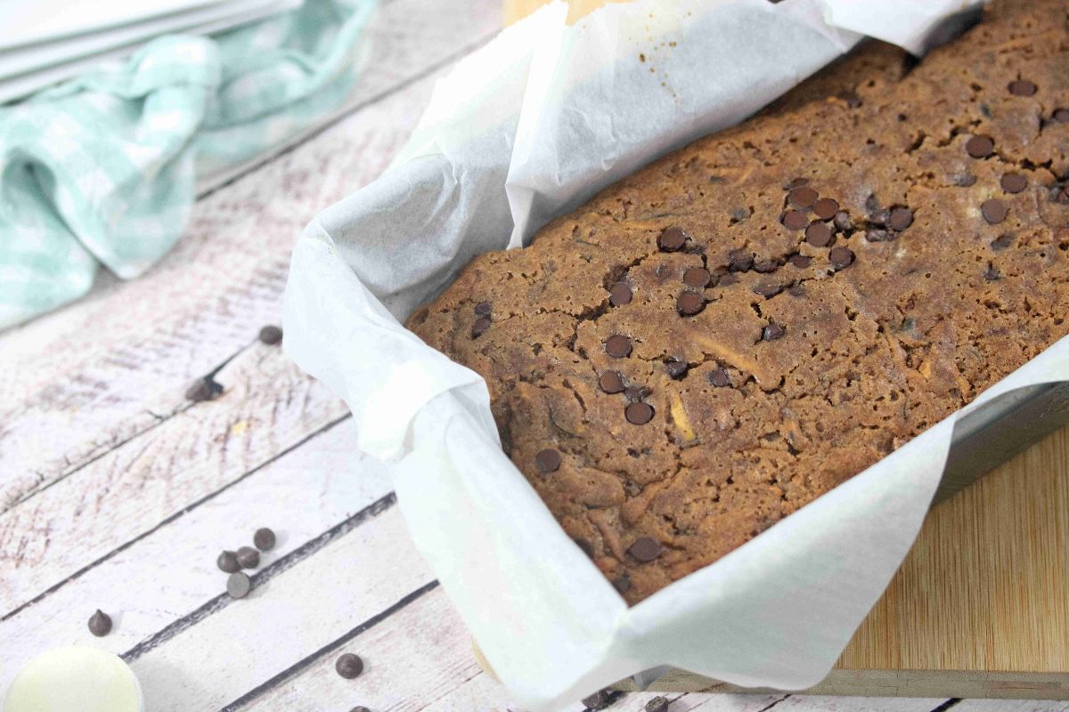 A loaf of dairy free chocolate chip banana bread in a parchment-lined baking pan on a light wooden surface—perfect for those who love indulgent lux desserts without the dairy.