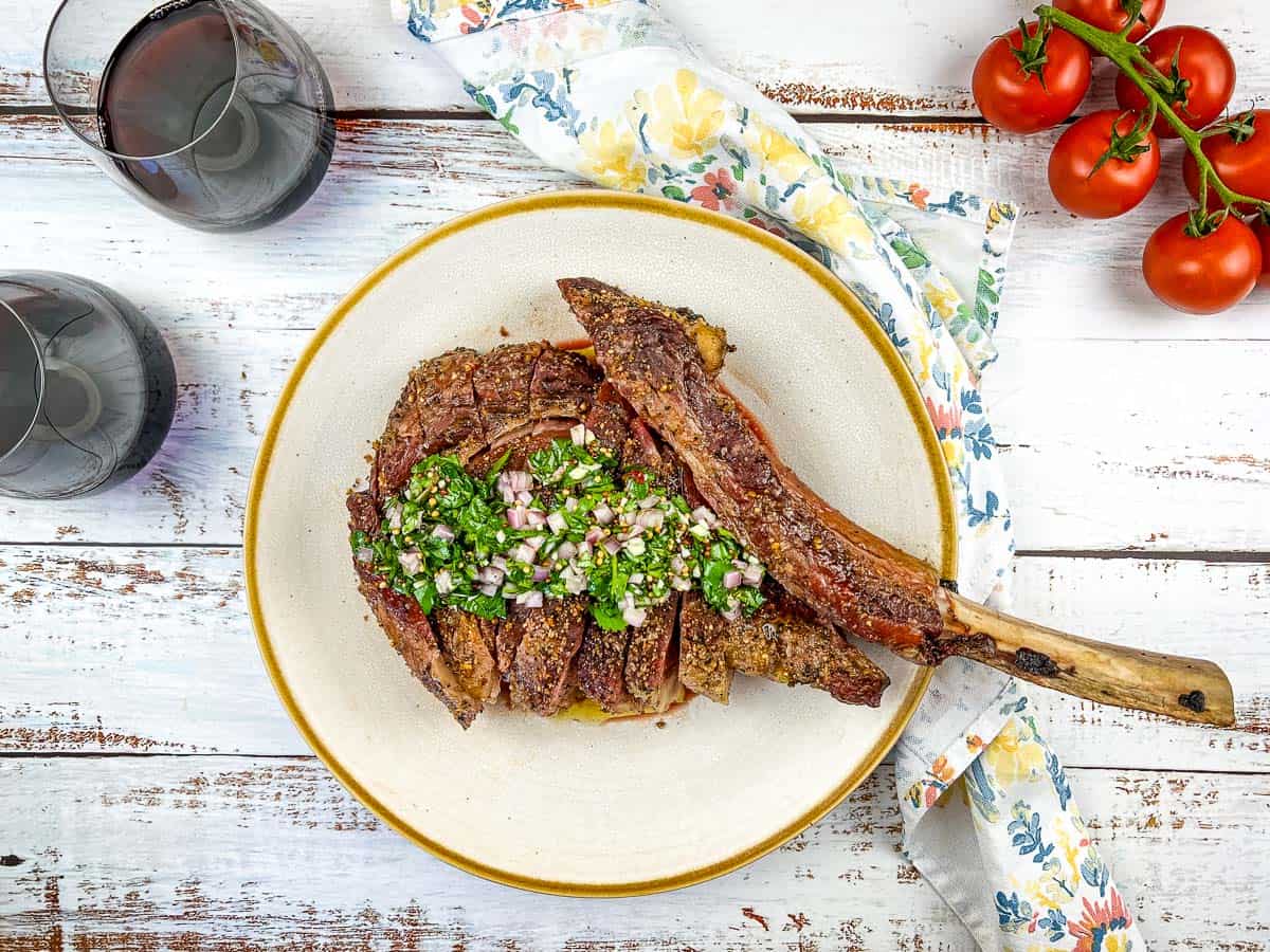 A sliced bone-in steak topped with chimichurri sauce on a plate, next to a floral napkin, two glasses of red wine, and vine tomatoes on a rustic white table—a perfect setting for fans of spicy recipes and heat and smoke flavors.