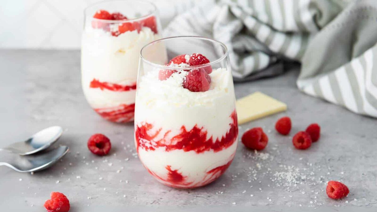 Two glasses filled with swirled layers of flavorful mousse and raspberry sauce, topped with raspberries and shredded white chocolate, with extra raspberries and spoons nearby on a gray surface. Perfect for fans of light desserts.