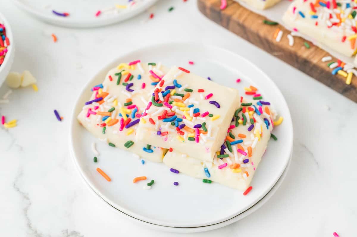 Two rectangular pieces of the best white chocolate fudge, topped with colorful rainbow sprinkles, are stacked on a white plate.
