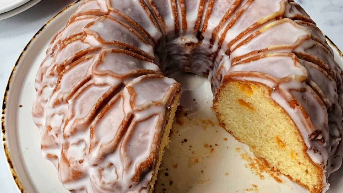 A Bundt cake with white glaze, partially sliced to reveal its moist, yellow interior dotted with fruit—perfect for celebration cakes—presented on a round ceramic plate.