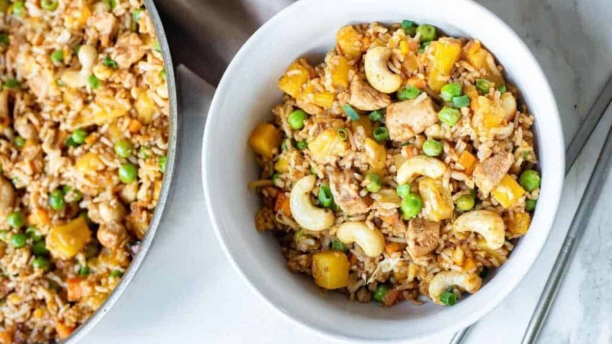 A bowl of gluten free fried rice with diced chicken, peas, carrots, cashews, and pineapple chunks sits next to a larger pan of this delicious dish—perfect for those seeking tasty gluten free recipes.