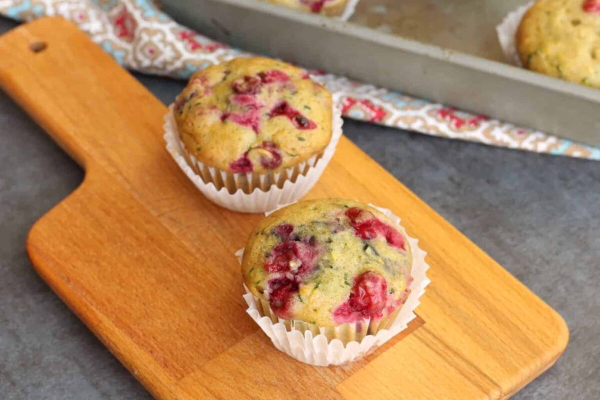 Two berry muffins in white paper liners sit on a wooden cutting board, offering cozy comfort. A metal baking tray and patterned cloth in the background complete this inviting scene—perfect inspiration for your favorite fall recipes.