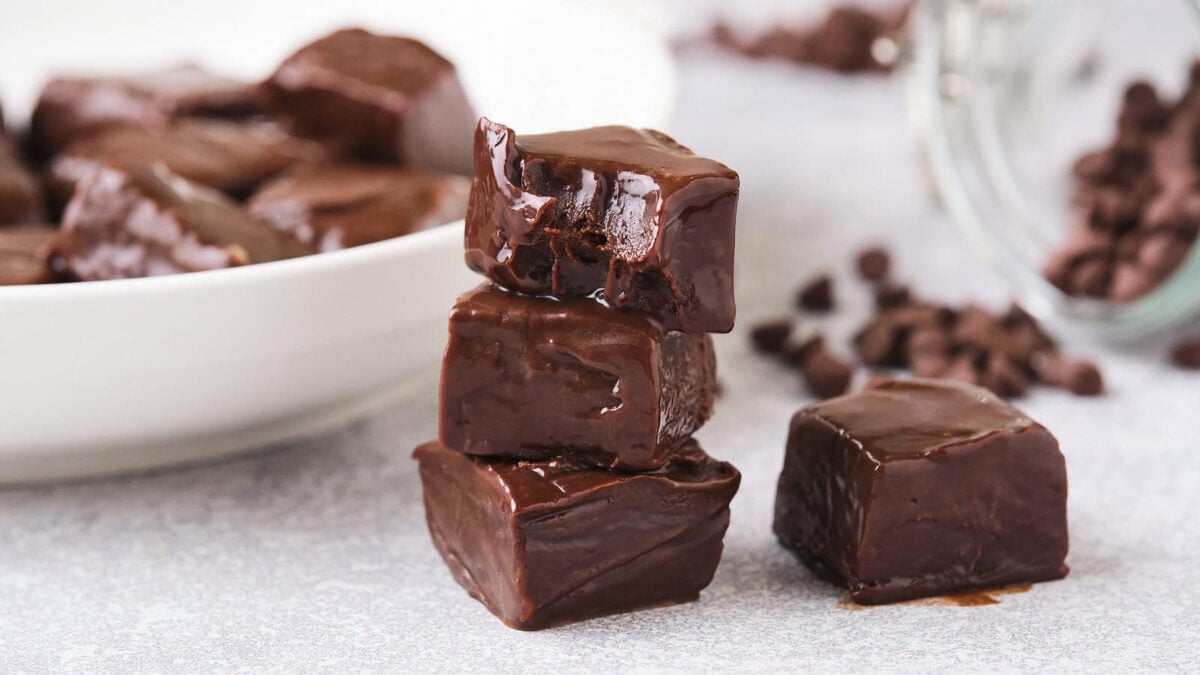 A stack of four glossy, square chocolate fudge pieces sits on a light surface; more fudge pieces and chocolate chips are in the background—perfect for those who love easy recipes.
