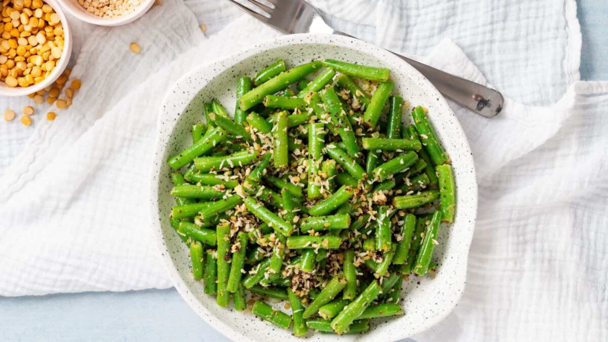 A white bowl filled with cooked green beans and tofu, topped with crushed nuts and seeds, placed on a white cloth with a fork and small bowls of ingredients nearby.