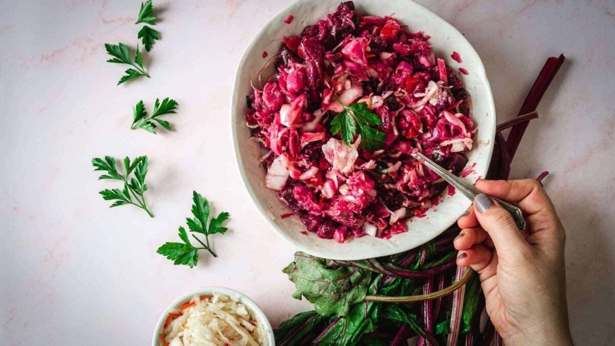 A hand holding a fork over a bowl of chopped beet and cabbage salad, with parsley garnish and beet greens—an inviting dish for fans of vegan recipes. A small bowl of sauerkraut on the side completes this vibrant plant-based meal.