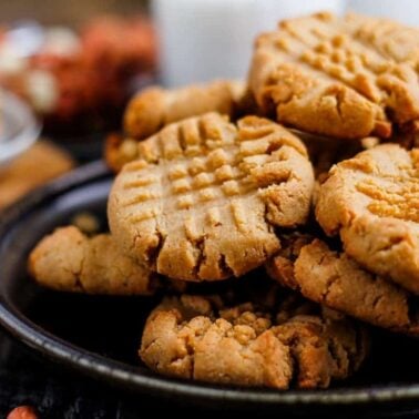 A plate of plant-based peanut butter cookies stacked on top of each other, with a crisscross pattern on each cookie. Peanuts and a glass of milk are blurred in the background.