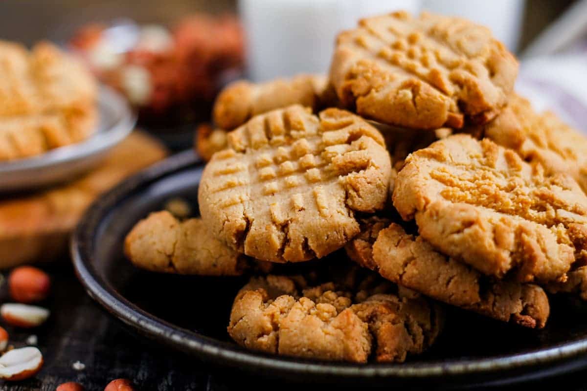A plate of plant-based peanut butter cookies stacked on top of each other, with a crisscross pattern on each cookie. Peanuts and a glass of milk are blurred in the background.