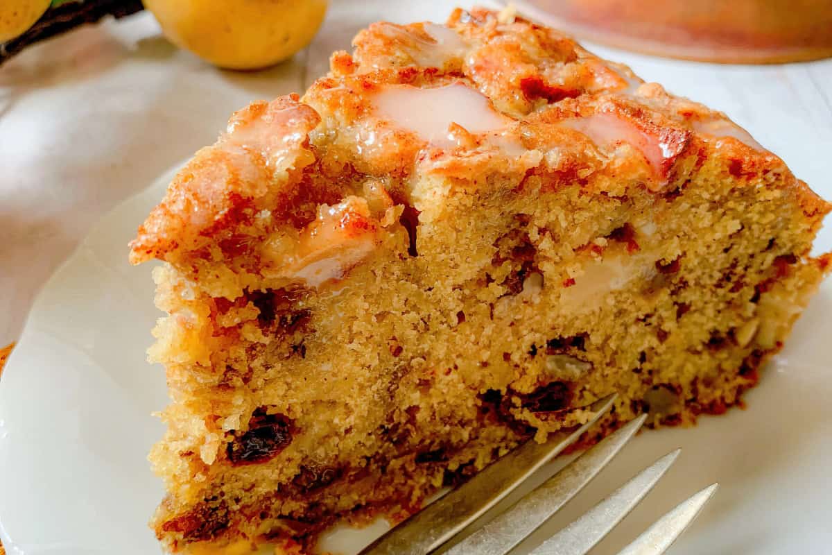 A close-up of a slice of impressive cake with fruit, nuts, and icing on top, sitting on a white plate beside a fork—a true showstopper cake for any occasion.
