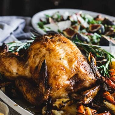 A roasted whole chicken on a platter with herbs and vegetables, accompanied by a salad in the background on a set dining table—perfect inspiration for easy dinner recipes and effortless entertaining.