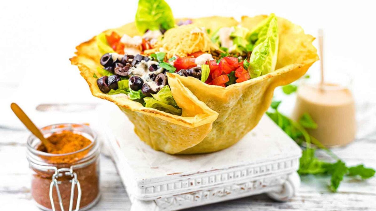 A taco salad served in a crispy tortilla bowl, topped with lettuce, tomatoes, black olives, cheese, and dressing. Perfect for fans of gluten free recipes, with a jar of seasoning on the side for extra flavor.