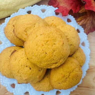 A plate of round, golden-brown cookies sits on a white decorative dish, surrounded by autumn leaves and a yellow cloth—perfect for cozy comfort and enjoying the warmth of fall recipes.