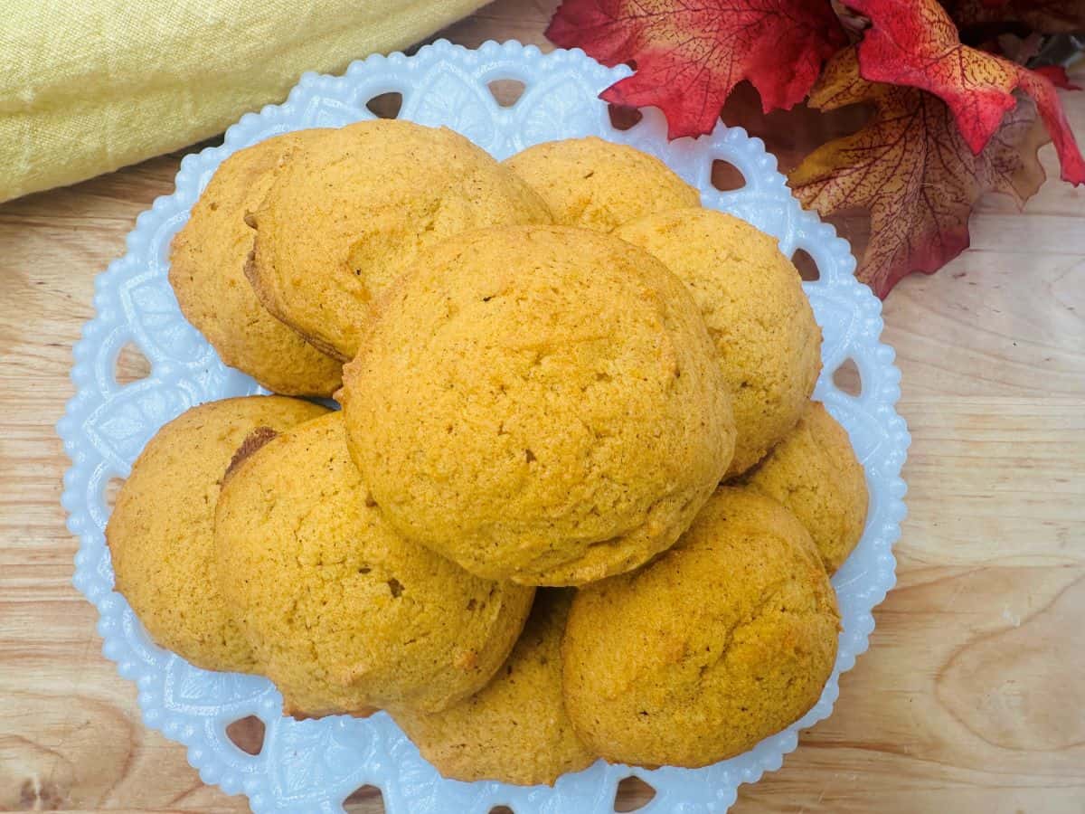 A plate of round, golden-brown cookies sits on a white decorative dish, surrounded by autumn leaves and a yellow cloth—perfect for cozy comfort and enjoying the warmth of fall recipes.